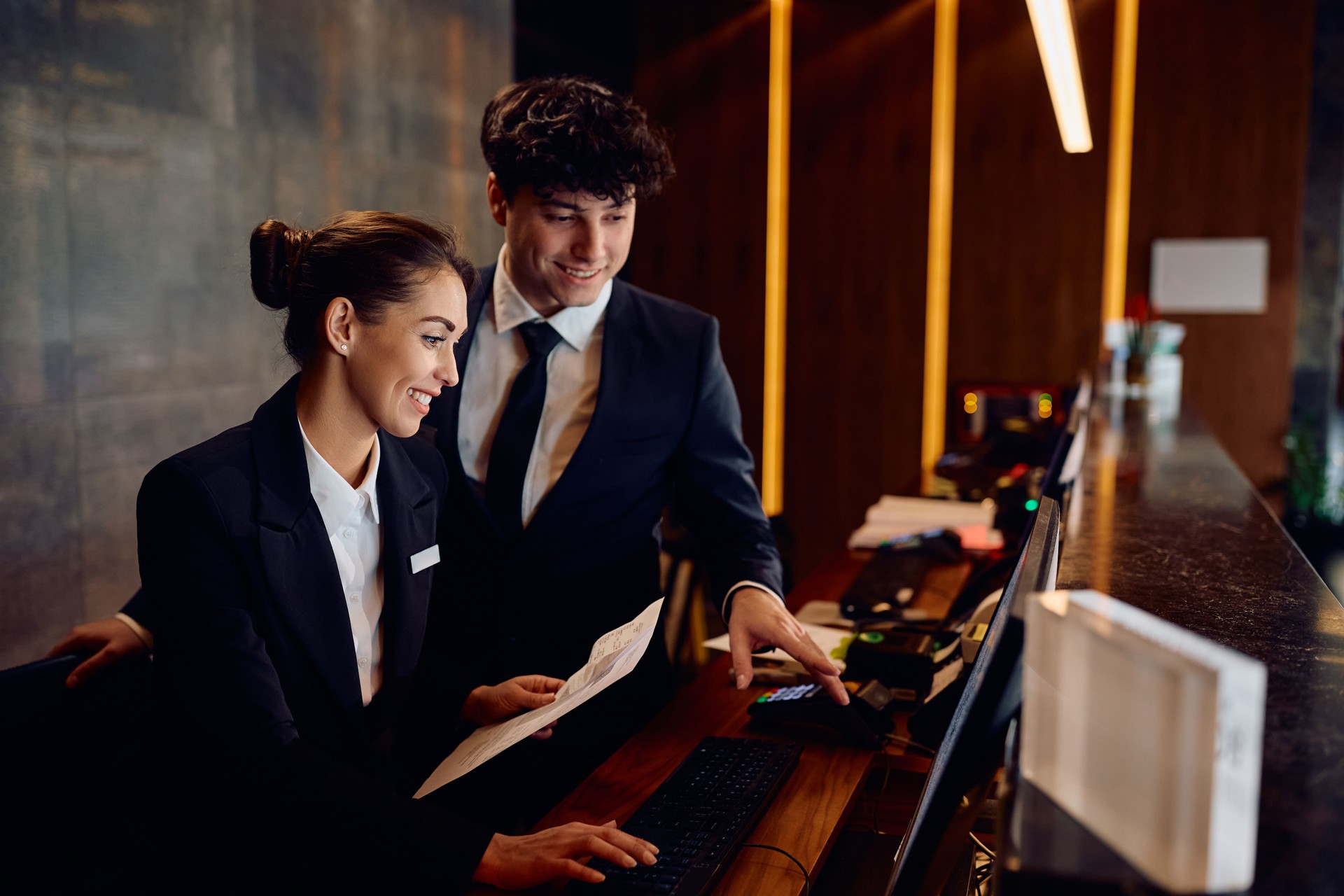 Happy receptionists cooperating while working on a computer at hotel front desk.